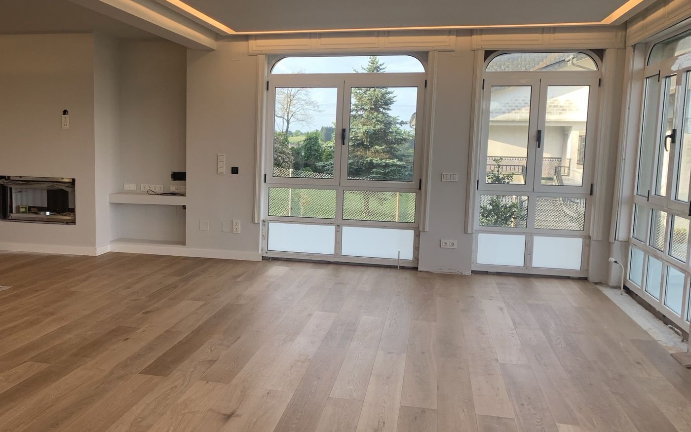 Open living room with oak plank floor, drywall ceiling with warm LED cove lighting and arched windows